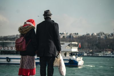 A couple stands near a river platform, waiting in winter clothing with boats in the background. Un couple se tient près d'une plateforme fluviale, attendant en vêtements d'hiver avec des bateaux en arrière-plan.