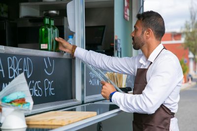 Side view of serious ethnic bearded male seller wearing apron standing near counter of food truck while preparing for job Vue de profil d'un vendeur barbu ethnique sérieux portant un tablier debout près du comptoir d'un food truck en préparation pour le travail