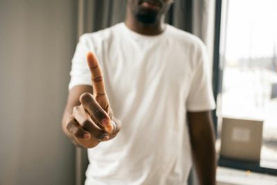 Homme africain américain anonyme en t-shirt blanc montrant un geste d'arrêt avec l'index pointé vers le haut dans une pièce lumineuse sur fond flou